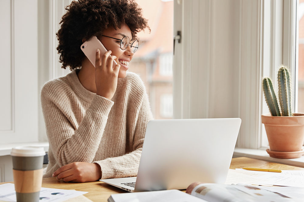 Horizontal shot of adorable cheerful dark skinned young lady calls to customer service, checks account balance, uses modern electronic gadgets, sits at desktop, looks with happy expression aside